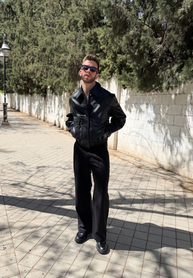 Man wearing black sunglasses, black leather jacket, and black pants stands on sunlit sidewalk with trees and white brick wall behind.