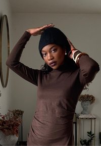 Haut marron à manches longues associé à un bonnet noir en maille. Accessoirisé avec plusieurs bracelets et boucles d'oreilles. Pièce avec des éléments décoratifs visibles.