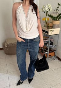 Femme portant un haut dos-nu blanc et un jean bleu, tenant un sac à main noir, debout à l'intérieur près de plantes et d'une étagère avec des objets décoratifs.