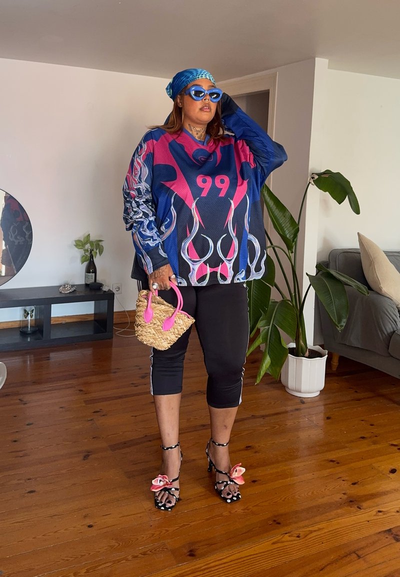 Personne portant une chemise oversize colorée avec le numéro 99, un pantalon noir coupé, des sandales avec des nœuds, un foulard bleu sur la tête, des lunettes de soleil et tenant un sac tressé.
