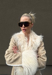 Mulher com cabelo loiro em um rabo de cavalo usa grandes óculos de sol e um casaco bege com colarinho e punhos de pele branca, de pé contra uma parede cinza texturizada.