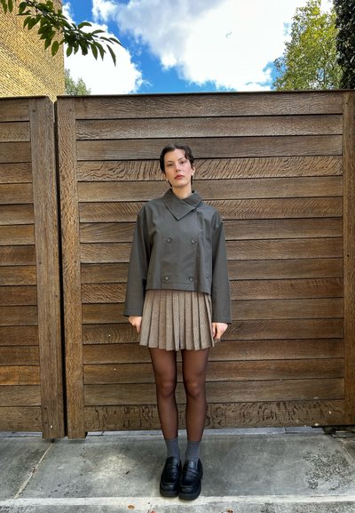 Chaqueta recortada en verde oliva con botones de doble botonadura, combinada con una falda a rayas marrón con pliegues y zapatos de plataforma negros.