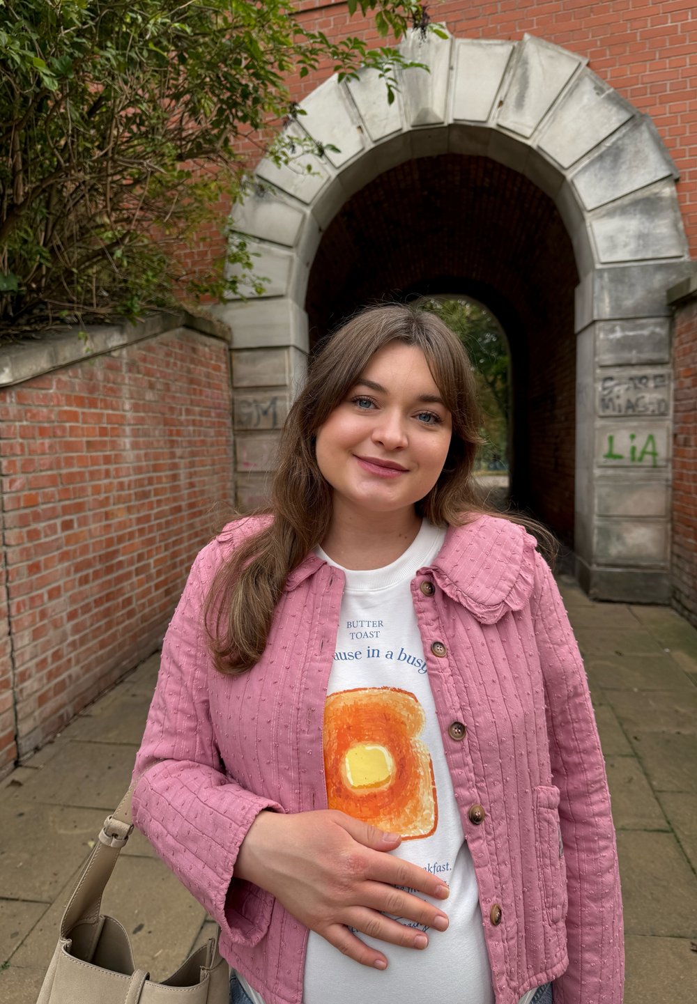Glimlachende zwangere vrouw in een roze jack staat bij een bakstenen poort, steunt met haar hand op haar buik en houdt een beige handtas vast op het geplaveide pad.