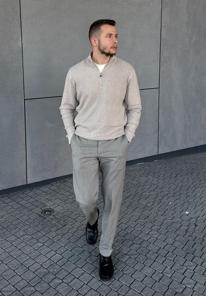 Man walking on tiled pavement, wearing light grey jumper, matching trousers, and black shoes, looking to his right against grey wall background.