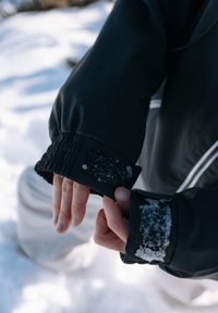 Personne ajustant la manche d'une veste noire avec de la neige sur les bandes Velcro, sol enneigé visible en arrière-plan.