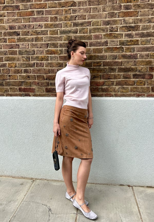 Helles rosa Kurzarm-Mock-Turtleneck, beige Wildleder Bleistiftrock mit floralem Schmuck, graue Ballerinas und eine kleine schwarze Handtasche.