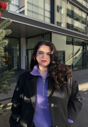 Abrigo negro de rayas finas sobre un top púrpura de cuello alto con cremallera. Cabello rizado oscuro, de pie al aire libre junto a un edificio de vidrio, con vegetación cercana.