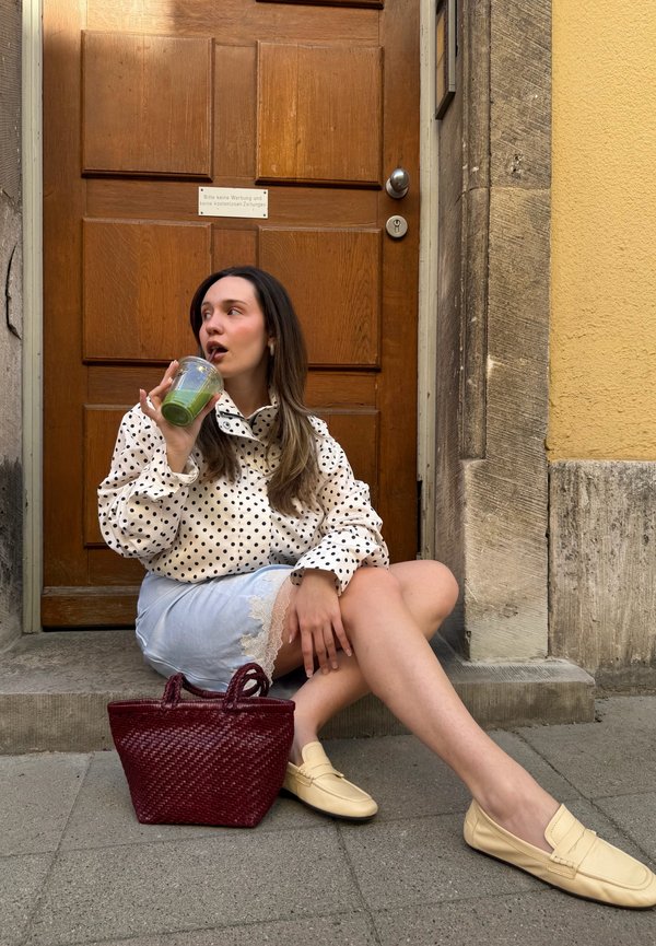 Kobieta w bluzce w groszki i spódnicy siedzi na stopniu, pije zielone smoothie, obok niej czerwona pleciona torba.