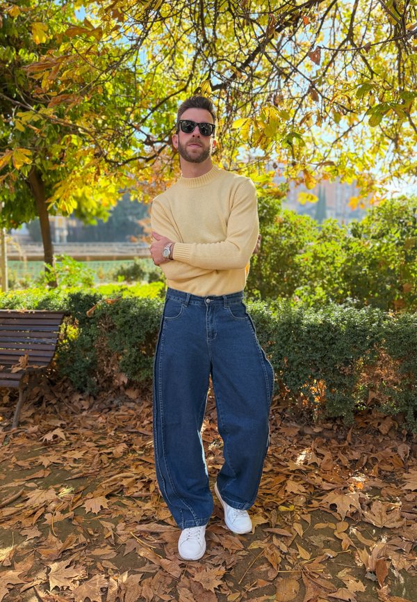 Homme portant des lunettes de soleil, un pull jaune et un jean bleu large, debout les bras croisés sous un arbre d'automne dans un parc avec des feuilles tombées.