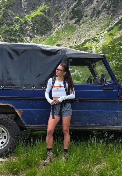 Vehículo todoterreno azul al lado de una persona con una camisa de manga larga blanca, pantalones cortos grises y botas de senderismo. Mochila con correas y acentos naranjas.