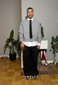 Grauer Strickpullover, weißes Hemd mit Kragen, schwarze Krawatte und lockere schwarze Hose. Accessoires umfassen eine Kette und eine Uhr. Neutraler Hintergrund mit Pflanzen.