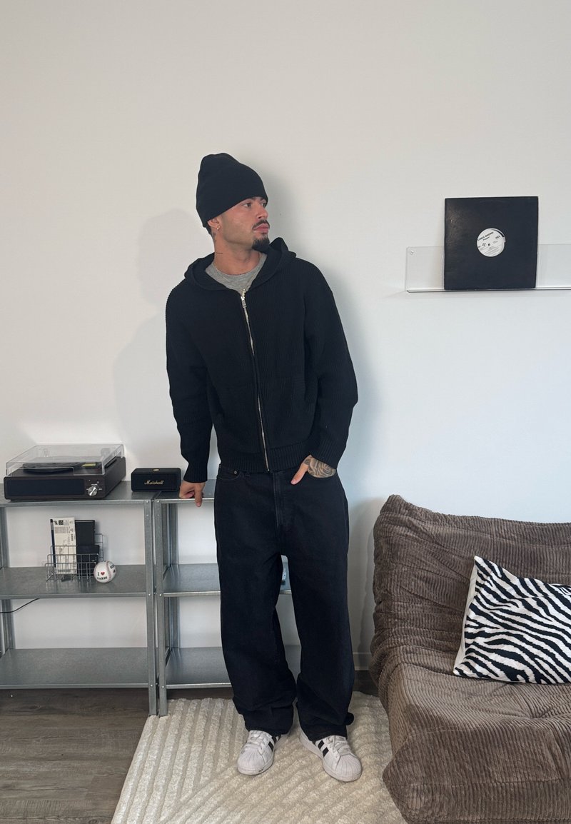 Sort lynlås-hættetrøje lagdelt over en grå trøje, kombineret med løse sorte jeans og hvide sneakers. Indeholder en mønstret zebra-pude i nærheden.