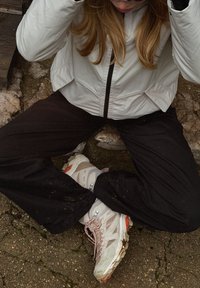 Hvid letvægtsjakke med lynlås, sorte bukser og off-white sneakers med røde accenter og synlige snørrebånd, der viser snavs på sålerne.