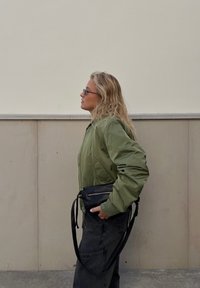 Olive green bomber jacket with a smooth finish, black zippered bag, and loose dark trousers, all set against a neutral wall backdrop.