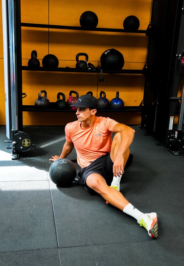 Joven con camiseta naranja y gorra negra sentado en el suelo del gimnasio junto a una pelota medicinal, con pesas rusas y equipo de ejercicio en estantes detrás de él.