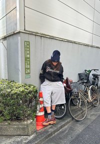 Personne portant un short ample, un sweat-shirt des Yankees et un bonnet, debout près de vélos et d'un cône de signalisation orange à un coin de trottoir en ville.