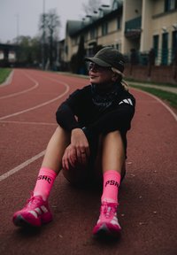 Atlétikai öltözék egy futópályán; fekete hosszú ujjú felsőt, "PSRC" feliratú rózsaszín zoknikat és élénk rózsaszín, fehér részletekkel díszített futócipőket tartalmaz.