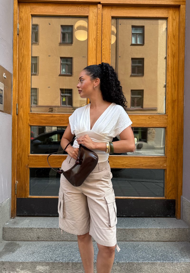 Femme aux cheveux bouclés, portant un haut blanc et un short cargo beige, tenant un sac à main marron foncé, debout près d'une porte en bois avec des vitres.