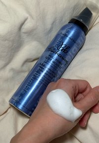 Mousse douce épaississante en forme pleine de Bumble and Bumble dans un récipient métallique bleu, avec de la mousse appliquée sur une main sur un fond de tissu clair.