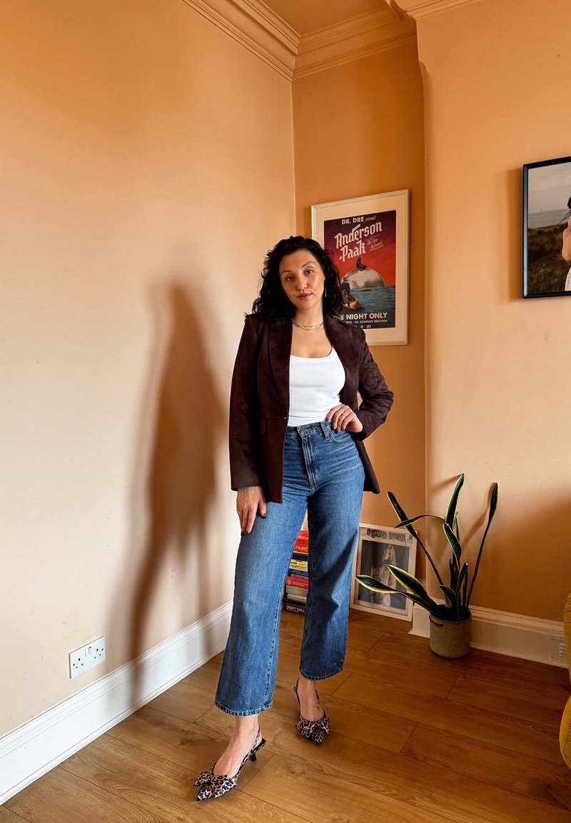 Brown blazer over a white tank top, paired with high-waisted blue jeans and leopard print heels; indoor setting with houseplant.