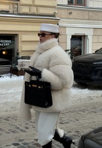 Personne portant un manteau blanc pelucheux, un chapeau assorti, des gants noirs et des lunettes de soleil, tenant un sac à main noir et deux gobelets de café à emporter dans une rue enneigée.