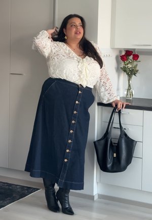 Mujer con blusa de encaje blanca y falda oscura abotonada está de pie en la cocina sosteniendo un bolso de cuero negro, con rosas rojas en un jarrón sobre la encimera.