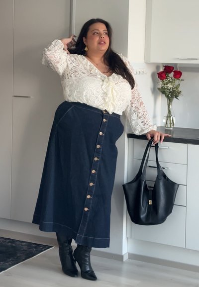 Mujer con blusa de encaje blanca y falda oscura abotonada está de pie en la cocina sosteniendo un bolso de cuero negro, con rosas rojas en un jarrón sobre la encimera.