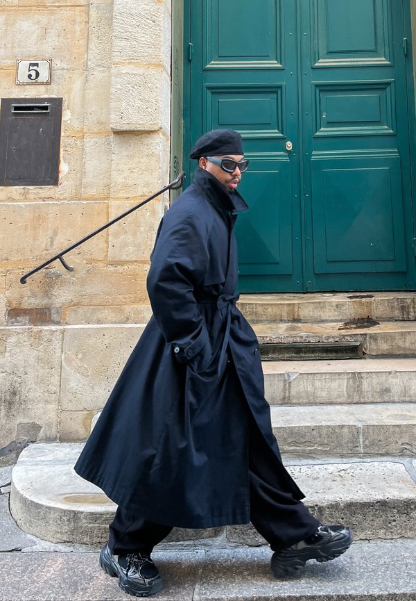 Pitkä musta trench-coat, jossa on solmittava vyötärö, päällä löysät mustat housut. Mustat kengät paksuilla pohjilla. Taustalla vihreät ovet ja kiviset portaat.