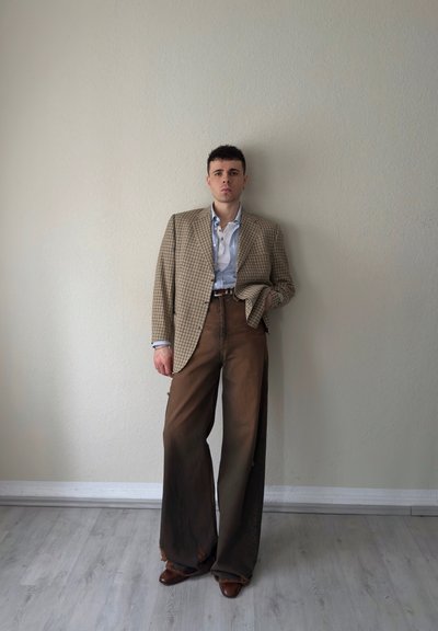 Joven de pie contra una pared beige, vistiendo un blazer marrón a cuadros oversize, camisa azul claro, pantalones anchos marrones y zapatos marrones.