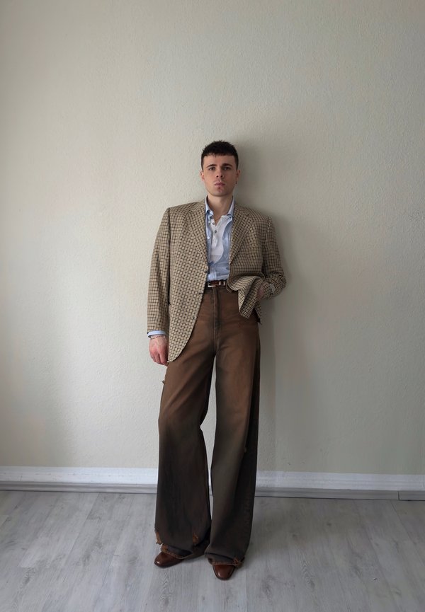 Jauns cilvēks stāv pret gaiši brūnu sienu, valkājot pārlielu brūnu rūtainu žaketi, gaiši zilu kreklu, platas brūnas bikses un brūnas kurpes.