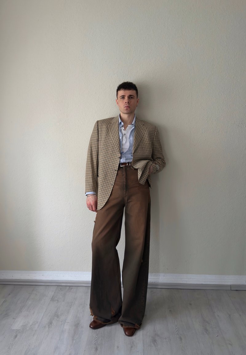 Mladić stoji uz bež zid noseći preveliki smeđi karirani sako, svijetloplavu košulju, široke smeđe hlače i smeđe cipele.