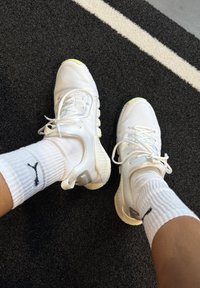 Ténis atléticos brancos com tecido texturizado e detalhes em verde, combinados com meias brancas caneladas com um logótipo preto, sobre uma superfície escura.