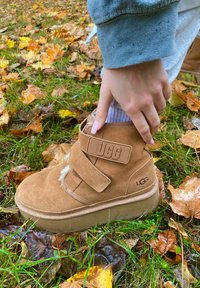 Rjavi nubuk ankel škornji z ježkom, mehka podloga vidna, gumijast podplat in UGG logo na strani, postavljeni med jesenskimi listi.