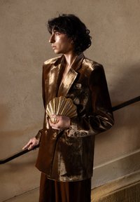 Personne en costume en velours marron brillant tenant une pochette dorée en forme de coquillage, debout sur un escalier avec la main gauche sur la rampe, regardant sur le côté.