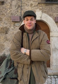 Jeune homme en veste marron et casquette, bras croisés, devant un bâtiment en pierre avec une porte en bois arquée portant le numéro 80.