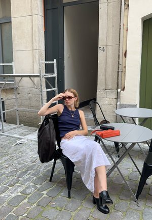 Una mujer en una camiseta sin mangas azul marino y falda blanca está sentada en una silla negra. Lleva gafas de sol y tiene una bolsa marrón, un bolso naranja y una mesa gris cerca.