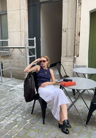 Una mujer en una camiseta sin mangas azul marino y falda blanca está sentada en una silla negra. Lleva gafas de sol y tiene una bolsa marrón, un bolso naranja y una mesa gris cerca.