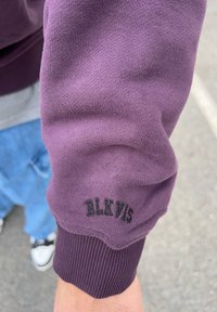 Tissu de sweat-shirt violet avec des poignets côtelés. Présente un logo "BLKVIS" brodé en noir sur la manche. Texture lisse et design décontracté.
