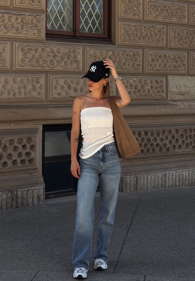 Top blanco sin tirantes, jeans rectos azul claro, gorra negra, bolso de hombro marrón, zapatillas blancas con acentos grises, de pie sobre un pavimento de piedra.