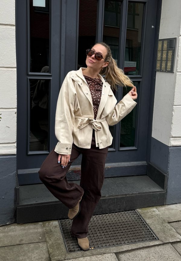 Kvinde med langt blondt hår iført solbriller, beige jakke, brun skjorte med hvide prikker, brune bukser og beige sko poserer foran en mørk dør.