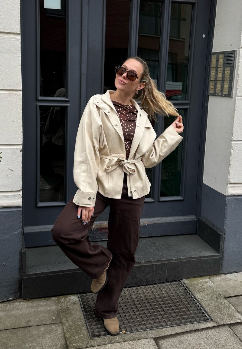 Frau mit langen blonden Haaren, die eine Sonnenbrille, eine beige Jacke, ein braunes gepunktetes Hemd, braune Hose und beige Schuhe trägt, posiert vor einer dunklen Tür.