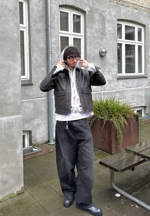 Zwarte lederen jas met twee voorzakken, laag over een lichtgrijze hoodie. Losse zwarte jeans, zwarte schoenen en een bril. Groene planten op de achtergrond.