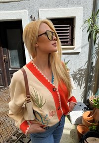 Megzta cardigan su oranžinėmis ir kreminėmis dalimis, puošta augalų siuvinėjimais, banguotais kraštais ir rudais mygtukais. Priedu yra šviesi rankinė.