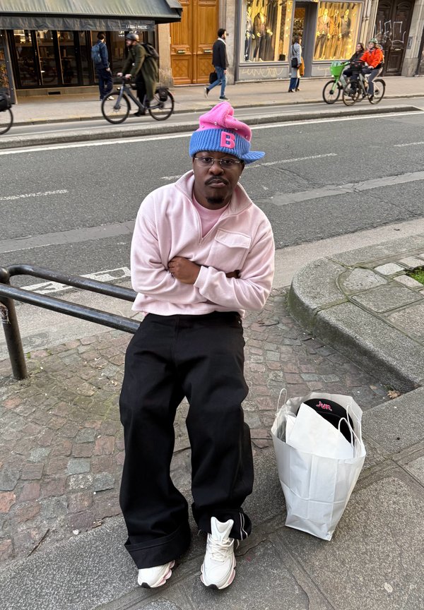 Mees roosas kampsunis ja kahvatoonilise sinise ja roosa mütsiga istub tänava äärekivil, tema kõrval on valge ostukott.