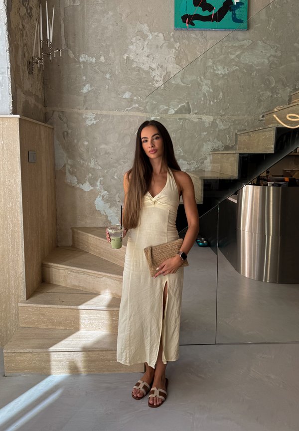 Una mujer con un vestido color crema sostiene una bebida verde y un bolso de mano tejido en un interior moderno, junto a una escalera de piedra.