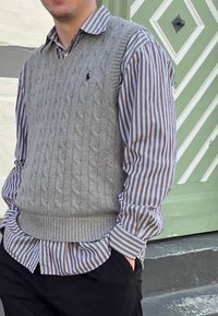 Hallikas nöörkootud vest pruunikas-valge triibulise kraega särgi peal, koos mustade pükstega. Vestil on ribaga äär ja pole nähtavaid logosid.