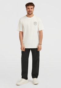 Hvid bomulds-t-shirt med korte ærmer, prydet med et cirkulært sort grafik på venstre bryst. Båret med sorte bukser og hvide sneakers.