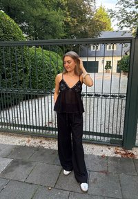 Top nero senza maniche con delicate ricami e orlo fluente, abbinato a pantaloni neri larghi e sneakers bianche. In piedi davanti a una recinzione.
