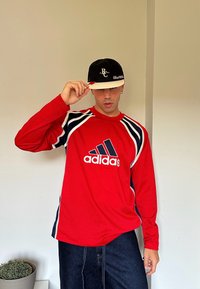 Rød Adidas langermet skjorte med hvite og marineblå striper, med logoen på forsiden. Modellen har på seg en svart caps med hvite detaljer.