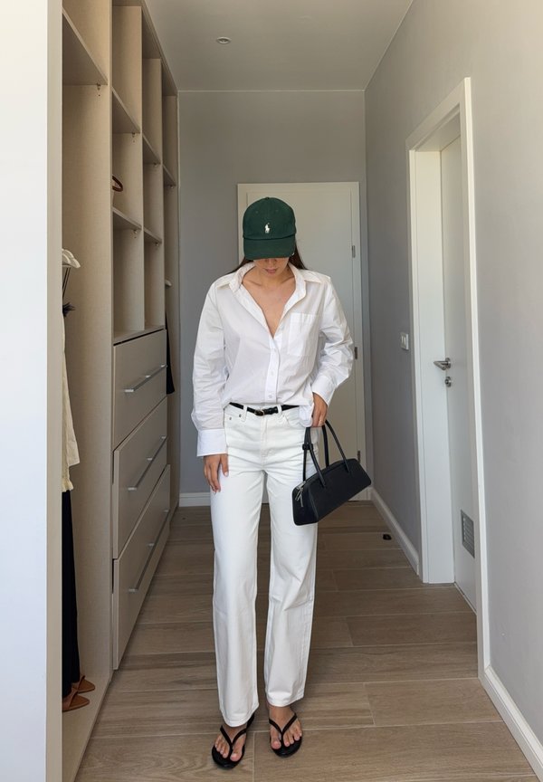 Mujer con camisa y pantalones blancos, gorra negra, sandalias y bolso, de pie en un pasillo con suelo de madera clara y paredes blancas.